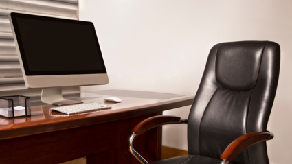 A leather office chair with varnished brown wood arm rests sit empty on front of a brown wooden desk. A laptop, keyboard, mouse and a stack of note paper sit on the desk.